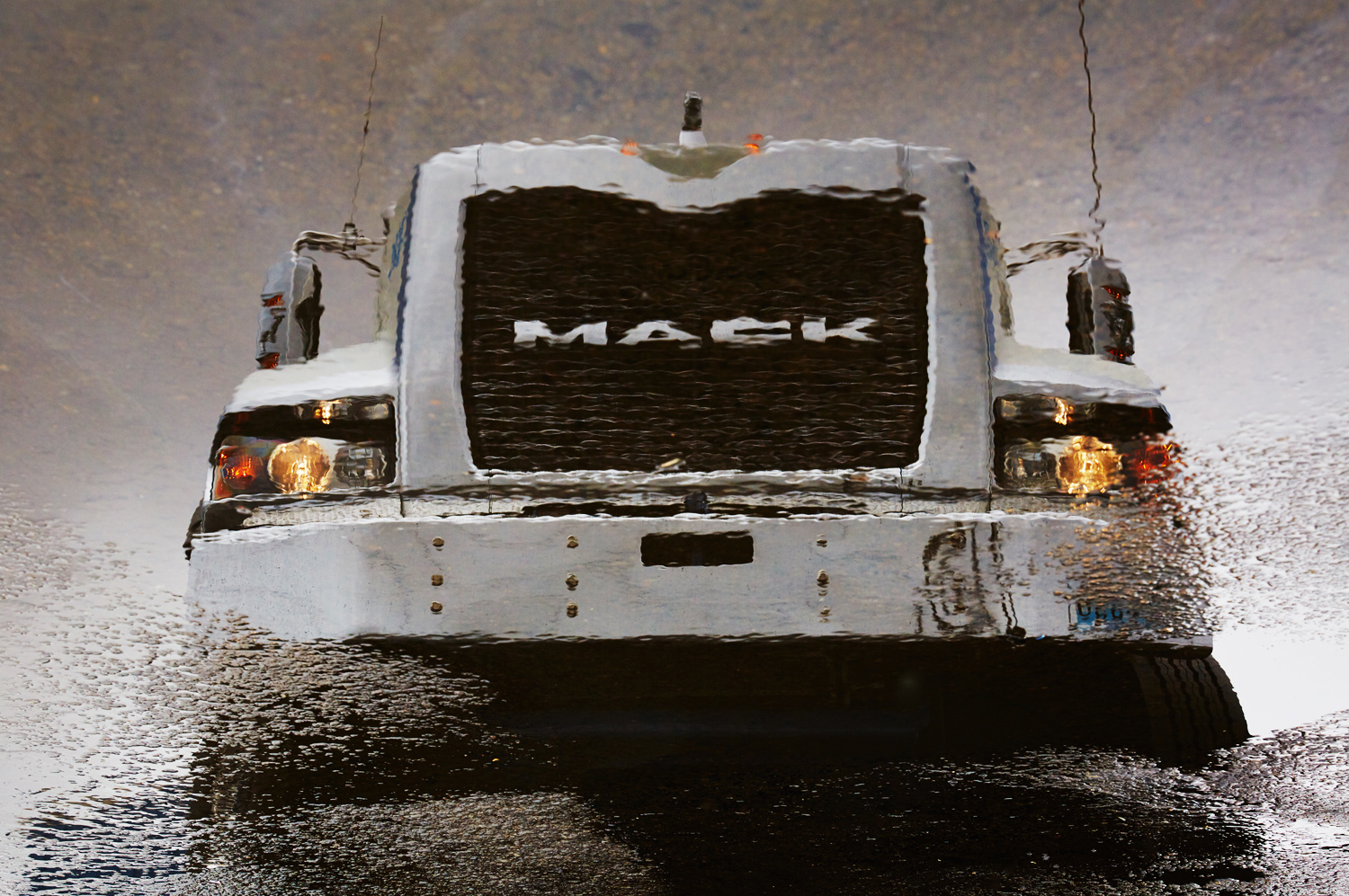 The front end of a Mack truck is reflected in a parking lot puddle.